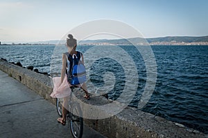 Young stylish woman in dress and with backpack is riding bicycle on promenade