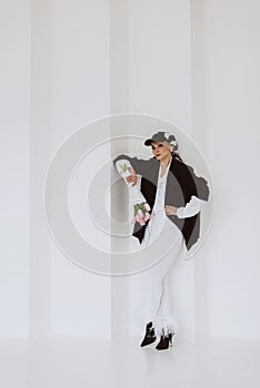 A young stylish woman in a fashionable outfit, a light white suit with feathers, a black jacket, a baseball cap and a bouquet of