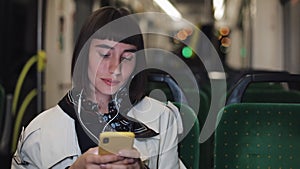 Young stylish woman in headphones listening to music and browsing on mobile phone riding in public transport. City