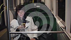 Young stylish woman in headphones listening to music and browsing on mobile phone riding in public transport. City