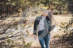 A young stylish woman standing in the middle of a forest or a park and looking directly into the camera and touching her hair with