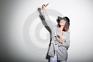 Young stylish woman using smartphone, indoor portrait over white. Fashion modern girl wearing grey jaket and black hat