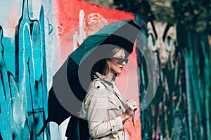 Young stylish woman wearing long beige coat, white boots, black hat, umbrella and backpack posing through the city streets.