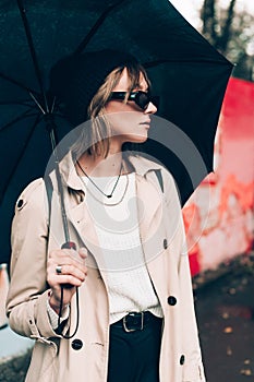 Young stylish woman wearing long beige coat, white boots, black hat, umbrella and backpack posing through the city streets.