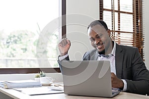 Young successful African American businessman uses laptop to work on white wooden table, expressing excitement in office
