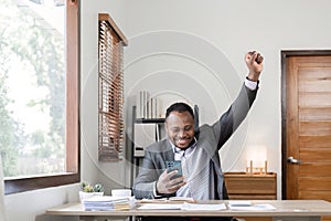 Young successful African American businessman uses laptop to work on white wooden table, expressing excitement in office