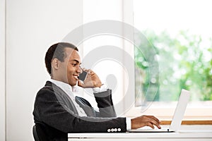 Young successful african businessman speaking on phone, typing laptop.