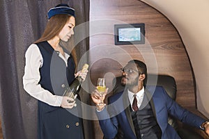 Young successful Afro-American businessman in glasses and a stewardess shows a bottle of wine in the cabin of a private