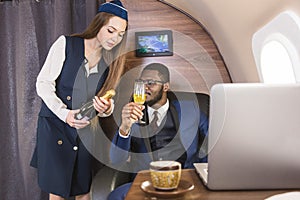 Young successful Afro-American businessman in glasses and a stewardess shows a bottle of wine in the cabin of a private