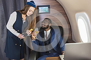 Young successful Afro-American businessman in glasses and a stewardess shows a bottle of wine in the cabin of a private