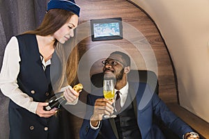 Young successful Afro-American businessman in glasses and a stewardess shows a bottle of wine in the cabin of a private
