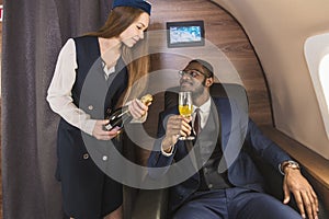 Young successful Afro-American businessman in glasses and a stewardess shows a bottle of wine in the cabin of a private