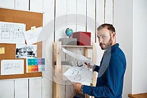 Young successful architector smiling, holding drawings, standing in office background.