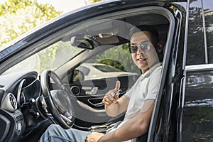 A young successful Asian businessman in sunglasses is sitting in an expensive modern car and smiling, looking at the camera
