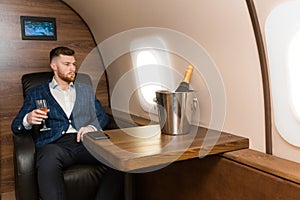 A young successful businessman in an expensive suit sits in the chair of a private jet with a glass of champagne in his