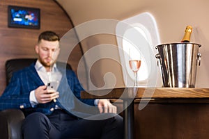 A young successful businessman in an expensive suit sits in the chair of a private jet with a glass of champagne in his