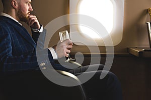 A young successful businessman in an expensive suit sits in the chair of a private jet with a glass of champagne in his