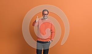 Young successful businessman in eyeglasses smiling and showing thumbs up sign on orange background