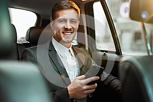 Young successful businessman holds his smartphone, working in the car on the way to his office, multitasking concept