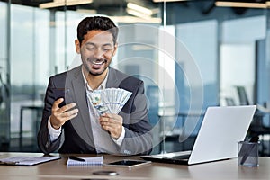 Young successful businessman inside office workplace holding phone and money cash US dollars in hands, man celebrating