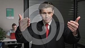 Young successful businessman in suit showing a come here sign. Man works on a computer in the background. 60 fps