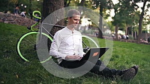 Young successful businessman in white shirt. Man sit on grass ground, work on laptop pc computer in city park on green