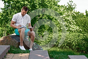 Young successful businessman in a white shirt. A man is sitting on the grass, working on a smartphone in a city park on a green