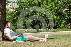 Young successful businessman in a white shirt. A man is sitting on the grass, working on a smartphone in a city park on a green