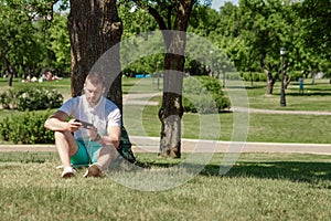 Young successful businessman in a white shirt. A man is sitting on the grass, working on a smartphone in a city park on a green
