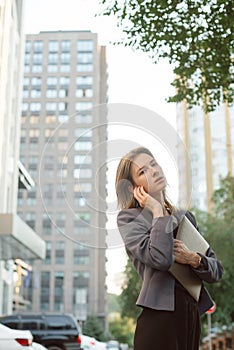 Young successful businesswoman in smart casual clothes in front of office buildings and skyscrapers. Woman holding modern laptop