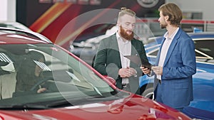 Young successful Caucasian businessman and confident car dealer talking in showroom as beautiful woman sitting on driver