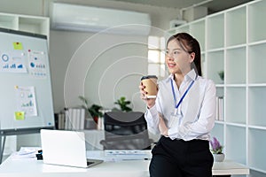 Young successful employee business woman hold craft paper cup coffee stand at desk with laptop pc computer at office