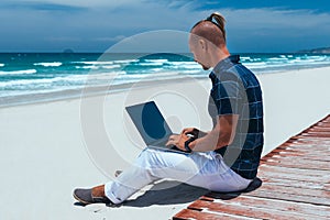 Young successful guy working using a laptop, sitting on the seashore on a sandy beach. Blogger, freelancer makes a content work