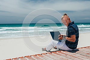 Young successful guy working using a laptop, sitting on the seashore on a sandy beach. Blogger, freelancer makes a content work