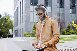 Young successful Hindu student studying online using laptop, man wearing headphones listening to music and online books