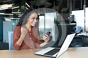 Young successful hispanic businesswoman working inside modern office, female worker using laptop, holding phone smiling