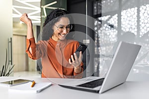 Young successful hispanic businesswoman working inside modern office, female worker using laptop, holding phone smiling