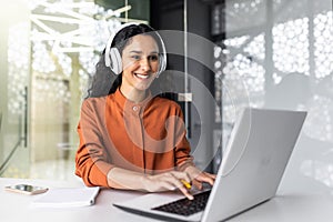 Young successful hispanic woman working in office with headphones, female programmer coding software on laptop smiling