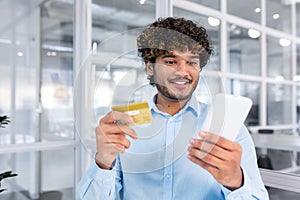 Young successful indian man close up in modern light office interior holding bank credit card and phone, businessman