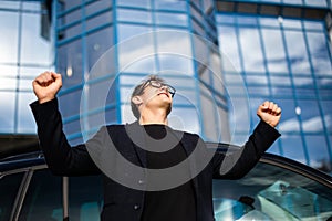 Young successful man, male executive businessman arms raised celebrating cheering shouting in front of a high rise office block in