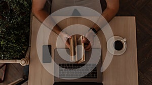 Young successful man sits at a table with a modern laptop with a cup of coffee with a mobile phone and holds a book in his hands.