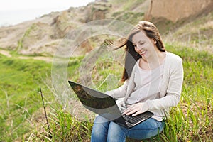 Young successful pretty smart business woman or student in light casual clothes sitting on grass using laptop in field