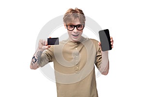 young successful red-haired caucasian guy dressed in a khaki short-sleeved shirt shows a credit card and a smartphone