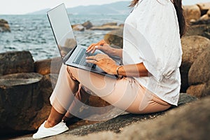 Young successful, smiling girl working using a laptop, sitting on a rocky seashore. traveling, blogger, freelancer, content plan,