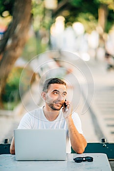 Young successful smiling smart man or student in casual shirt, glasses sitting at table, talking on mobile phone in city park usin