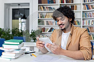 Young successful student studying sitting inside university academic library, man smiling holding tablet computer