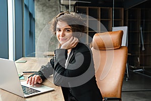 Young successful woman in business suit, sitting in her office corporation, lady boss working on laptop near window