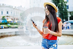 Young summer beauty woman with phone chating or surf in internet against fountain in the street