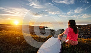 Young surfer girl sitting at the ocean coast near her surfboard, watching the sunset and enjoy the nature while deep breathing