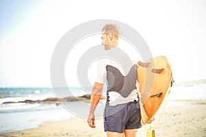 Young surfer holding his surfboard looking the waves for surfing - Handsome man standing on the beach at sunset training to surf